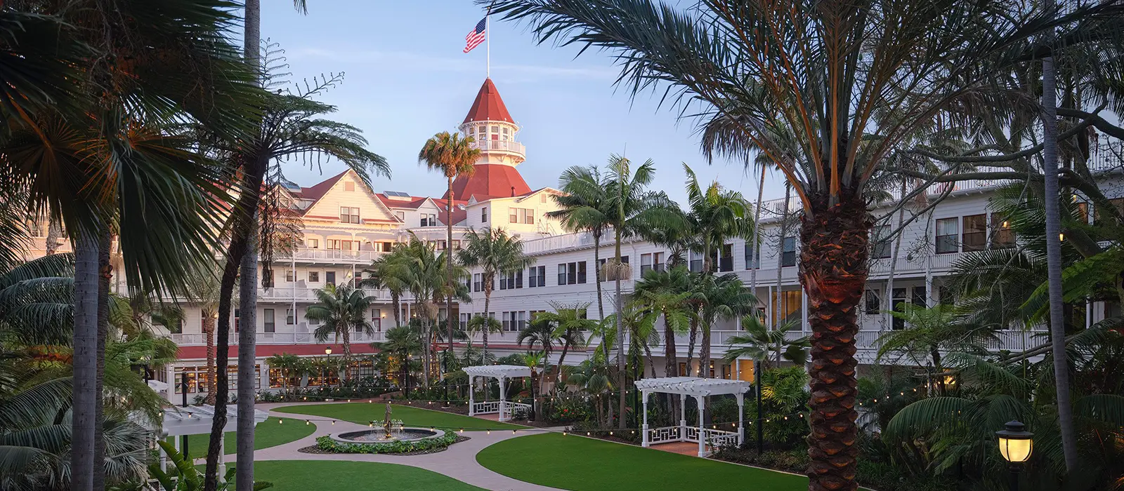Hotel del Coronado courtyard with palm trees, gardens, and the iconic red turret at sunset.